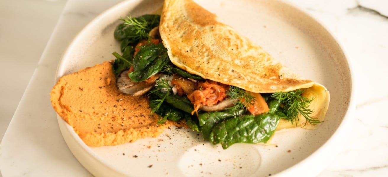 Savory omelette filled with leafy greens, sautéed mushrooms, and tomatoes, served with a side of roasted red pepper hummus on a minimalist plate.